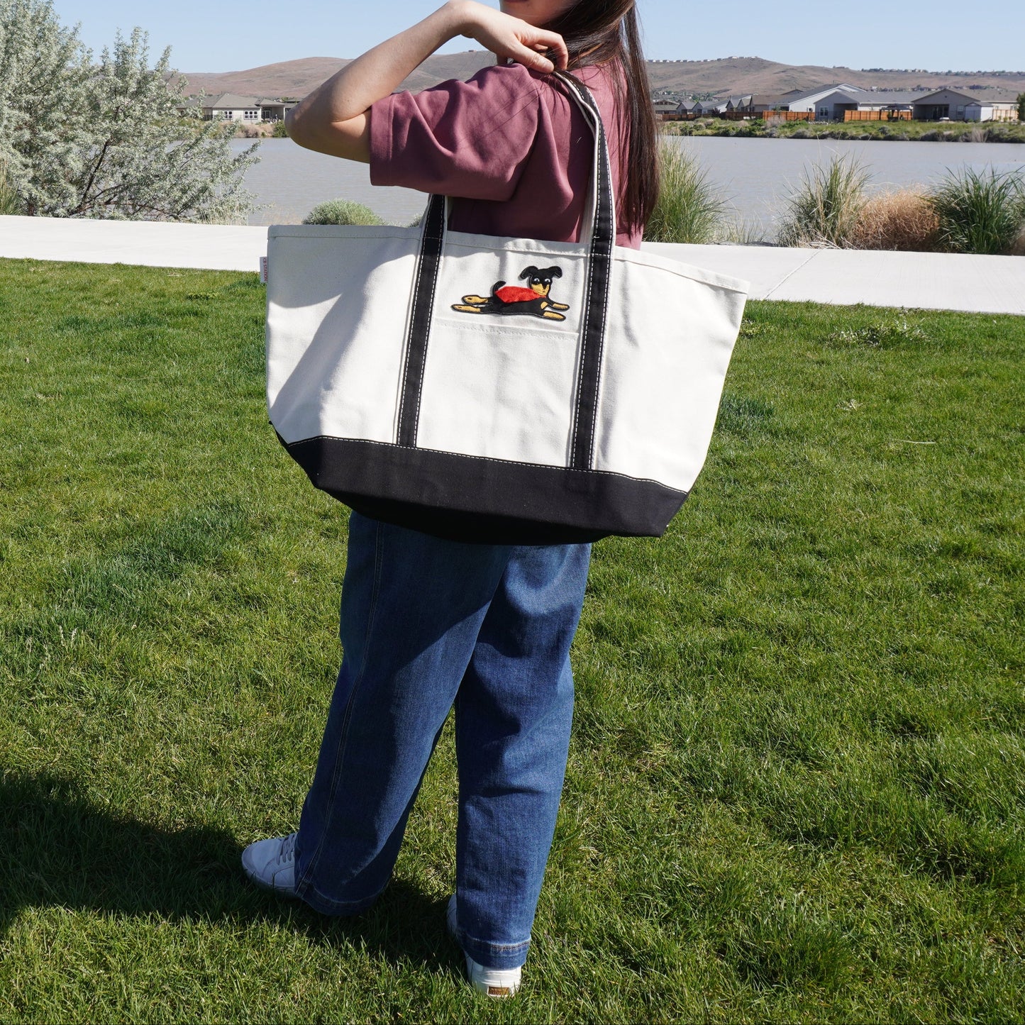 A woman standing on a grassy lawn outdoors, carrying a large off-white canvas tote bag with dark trim and handles. The tote bag has a small embroidered graphic on the front.